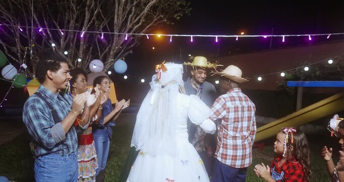 A joyful group of people celebrates a traditional Brazilian Festa Junina mock wedding ceremony at night, featuring a bride and groom in typical caipira costumes under colorful festive lights