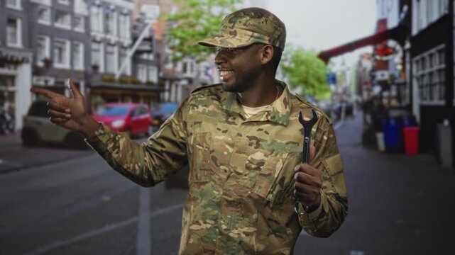 Soldier man in camouflage points finger while holding wrench and smiling on a busy street near parked cars and buildings; pride duty resilience camaraderie.
