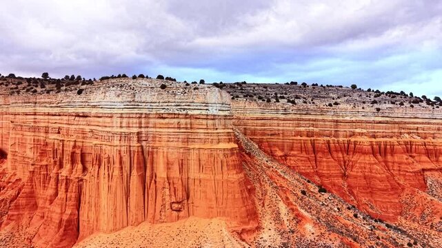 Aerial view of tall eroded red sandstone rock formations with horizontal white sedimentary layers on top under overcast weather. Red sandstone cliffs under cloudy sky.