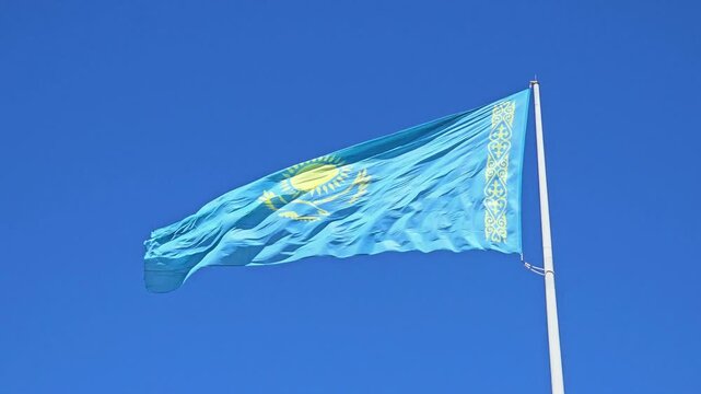 The flag of Kazakhstan flutters in the wind against a clear blue sky. A close-up of the national flag of the Republic of Kazakhstan against a clear sky.