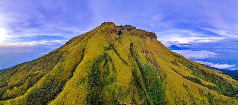 Aerial view of the lush green slopes and volcanic peak of Mount Sumbing under a blue sky with a sea of clouds in Temanggung, Central Java, Indonesia.