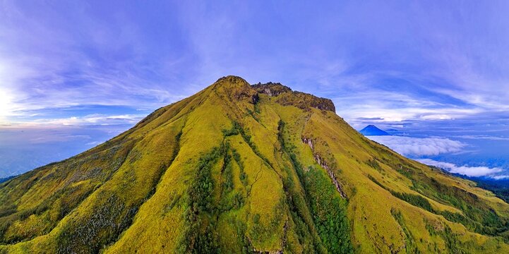 Aerial view of the lush green slopes and rugged peak of Mount Sumbing under a clear blue sky with a sea of clouds in the distance Temanggung, Central Java, Indonesia.