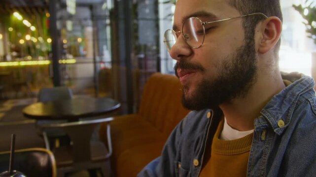 Close up shot of young biracial man with beard and glasses appreciating and taking bite of cheeseburger at table in modern restaurant