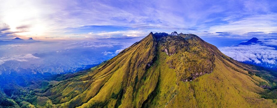 Aerial view of Mount Sumbing with its steep green slopes and rocky peak above a sea of clouds during sunrise in Temanggung, Central Java, Indonesia.