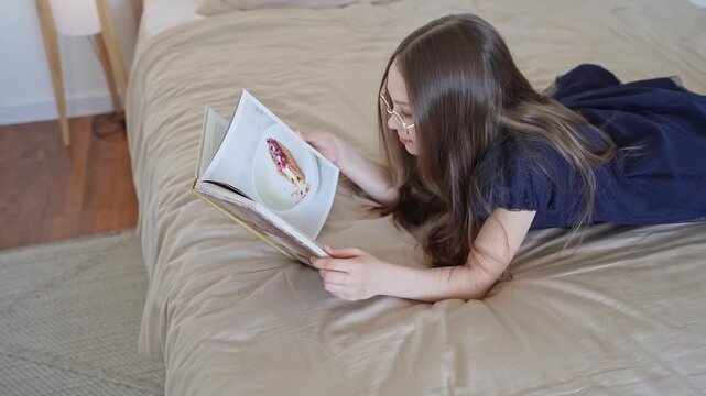 Girl with wavy hair reading hardcover book while lying on bed with soft blanket. Book, bed, wavy hair, blanket, girl, reading.