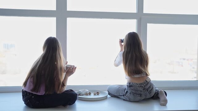 Two girls sitting quietly on windowsill looking at city skyline in peaceful daylight. Sitting, windowsill, girls, city, daylight, quiet.