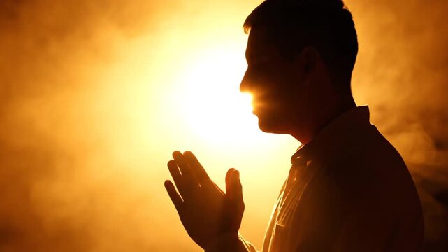 Profile silhouette of a person with hands folded in a prayer gesture, backlit with rim lighting against a solid black background, showing quiet devotion, faith, and spirituality.