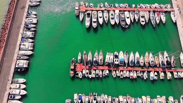 Aerial shot of various white powerboats and sailboats docked at red piers in turquoise water. Rows of boats in marina.