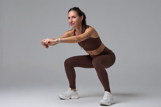 Fit woman doing squatting exercise looking at camera on gray background