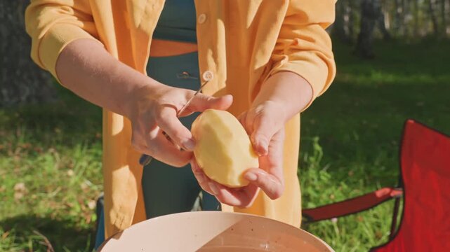 Camper finishing potato prep at campsite. Hands rinsing and trimming tuber over bowl, sunlight through trees, red folding chair on grassy clearing, steady motions preparing vegetables for campfire