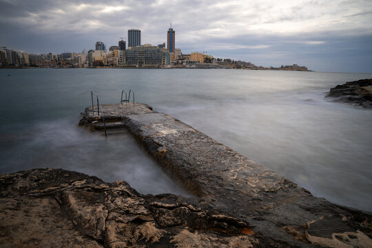 Long Exposure of Concrete Swimming Jetty on Rocky Coast with St Julians Skyline and Portomaso Tower in Background, Malta