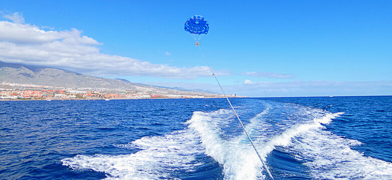 Colorful parasail soaring behind motorboat across Atlantic Ocean near Tenerife coast with mountain panorama and bright blue sky. Dynamic summer tourism activity with sea spray and tropical