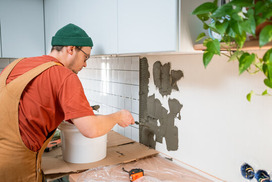 Man in overalls and beanie applying tile adhesive to a kitchen wall for a new backsplash, performing diy home renovation and improvement work to save money and update the interior
