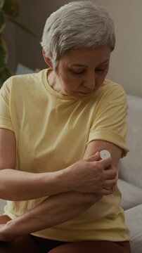Vertical dolly in shot of senior woman adjusting adhesive glucose sensor on upper arm while during routine diabetes management at home, slow motion
