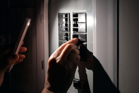 Closeup of person using smartphone flashlight while switching electrical circuit breakers in dark home. Residential electricity, power outage, emergency, troubleshooting, or electrical safety concept