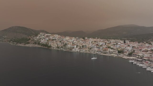 Drone flying backward revealing Ermioni coastal town and marina while sailing yacht slowly leaves the harbor under cinematic dusty orange sky