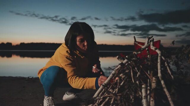 Evening camp setup by lake, camper arranging stick teepee and kindling, methodical handson prep for fire, red chair and calm water backdrop, practical survival skill in moody twilight