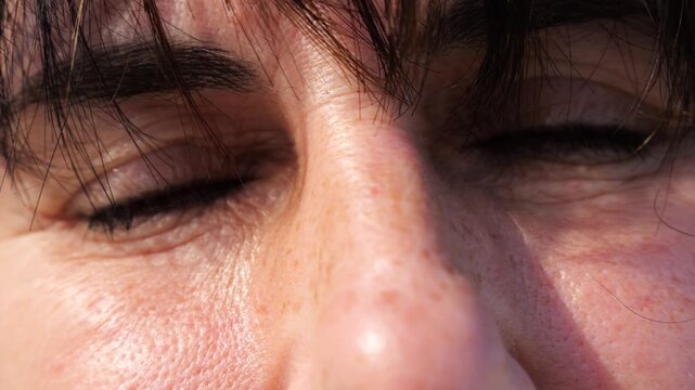 Tired blue eyes of a female blinking and looking into camera outdoor. Face of adult woman illuminating by bright rays of sun. Upset facial expression of lady. Close up Slow motion