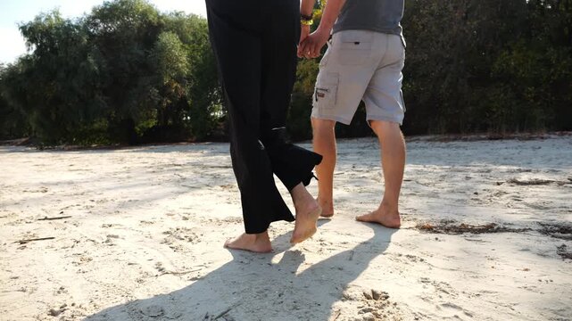 Happy pair going along coast at sunny day. Romantic couple holding hands of each other and walking on sandy beach. Lovers spending time together at resort. Concept of summer vacation or holiday
