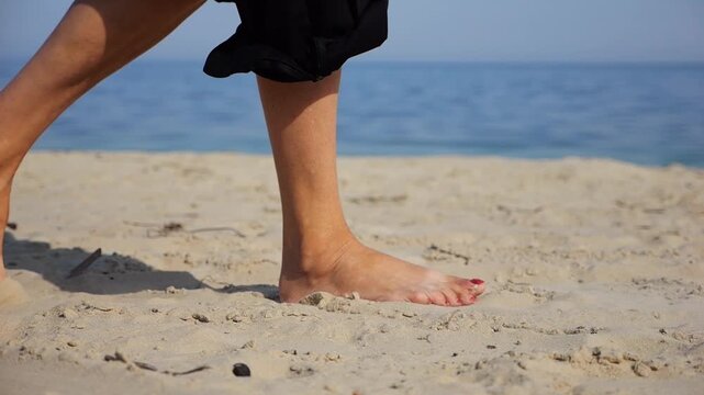 Female feet walking on sand at the beach with seascape at background. Legs of adult woman stepping along sea shore. Barefoot lady strolling near ocean coast. Concept of summer vacation. Low view