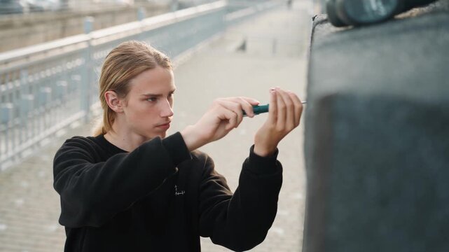 White man adjusting metal bracket on bridge parapet, urban waterfront walkway in background, concentrated hands using screwdriver, braided hair and black sweater convey careful craftsmanship