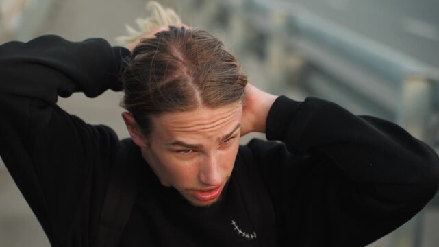 Youth hairstyle scene. Teenage boy securing his hair into bun amidst urban surroundings at dusk. Young man with cornrow hairstyle adjusting his hair bun overpass cityscape evening atmosphere