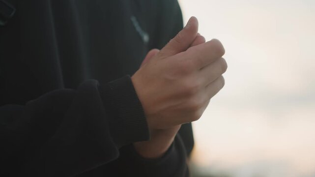 Sunset hands soothing palm rubs, person in dark sweater calms with slow finger motions, soft sky silhouette and warm ambient light, reflective rooftop mood, gestures alternate between clapping,