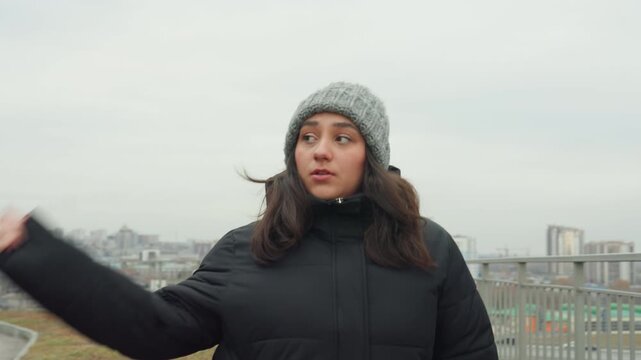 Woman dancing on urban overlook wearing beanie and puffer coat, playful arm swings and steady steps along path, city panorama behind, closeup expressions alternating with wide shots, rhythmic practice