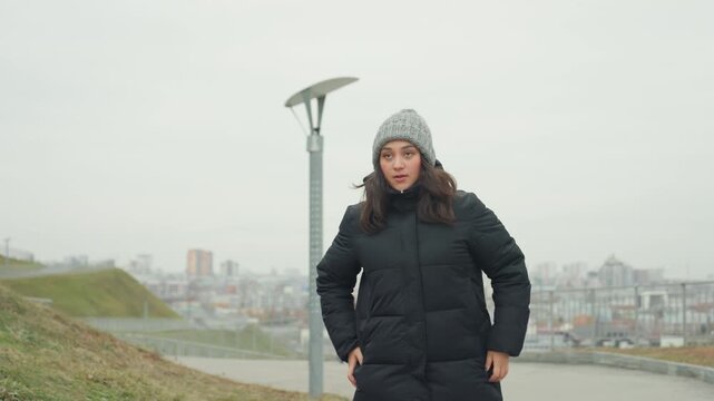 White woman warming up with kick, energetic rehearsal on hill slope in parka and knit hat, expressive footwork and quick stride, city skyline backdrop, casual outdoor practice, visible breath in cold