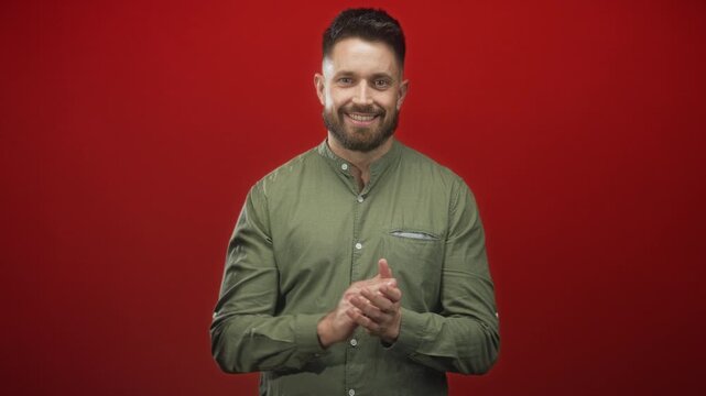 Man claps hands and smiles at camera in studio against red backdrop wearing green button shirt and beard; approval.