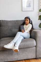 Obraz premium Asian child girl sitting on gray sofa at home wearing headphones holding smartphone, copy space