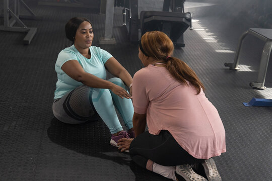 Two women performing sit-ups on rubber floor, turquoise woman holding feet, pink partner kneeling