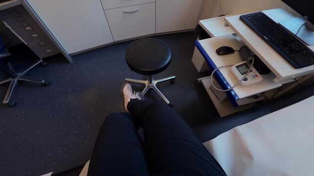 Patient perspective while sitting on an examination table in a modern doctor's office in Germany. The room is equipped with a sink, clinical supplies, and computer workstations for diagnostics.