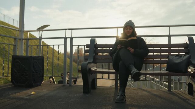 Sunlit bench closeup white woman dancing, smiling while seated with tablet knit hat and casual coat, bag at side, relaxed commuter vibe with soft backlight and warm glow subtle foot movement