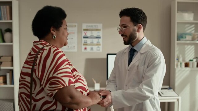 Medium shot of Middle Eastern male doctor greeting Black female patient in modern office with desktop computer before medical consultation