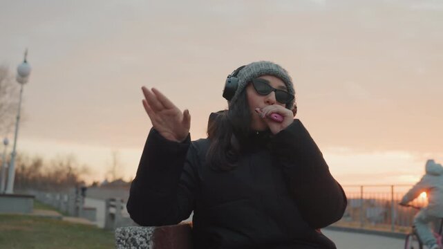 Woman on bench dancing freestyle to music at sunset, animated hand choreography, beanie and headphones, sunglasses reflecting golden sky, joyful grin, park walkway and city skyline, energetic