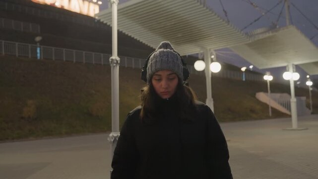 Blurry foreground passerby reveals female listener by lamplight, headphones and beanie in sharp focus, quiet rhythm implied against marquee lights and urban architecture, introspective performer