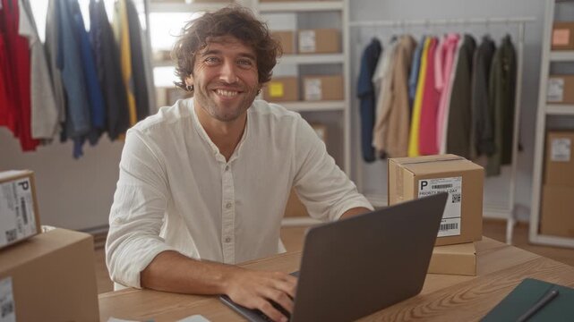 Man works on laptop amid cardboard shipping boxes and gives thumbs up with his hand while seated at a packing table in a studio; small business optimism.