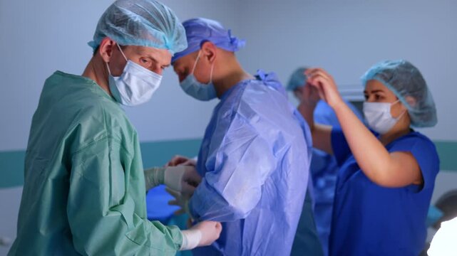 Medical staff helping the surgeon to get dressed for the surgery. Female nurse ties the robe and male assistant helps to put on the gloves.