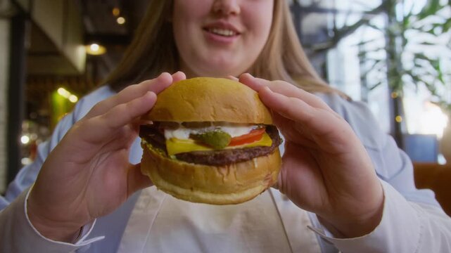 Low angle shot of young woman holding burger with two hands and anticipating taking bite in modern fast food cafe
