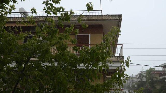 Building with balconies and closed roller shutters on windows during rain. Cloudy video of downpour and water running off roofs with trees swaying in wind. Resort town in off season.