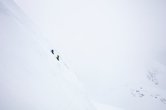 Two climbers ascending a steep snow covered ridge deep in the mountain clouds.