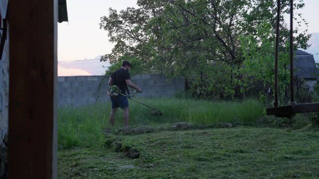 Man Using String Trimmer to Cut Tall Grass in Rustic Backyard at Dusk