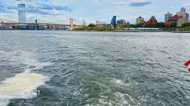 Boat with American flag on the pole moves by the river. The Manhattan Bridge, the Brooklyn Bridge and New York skyline in the backdrop.