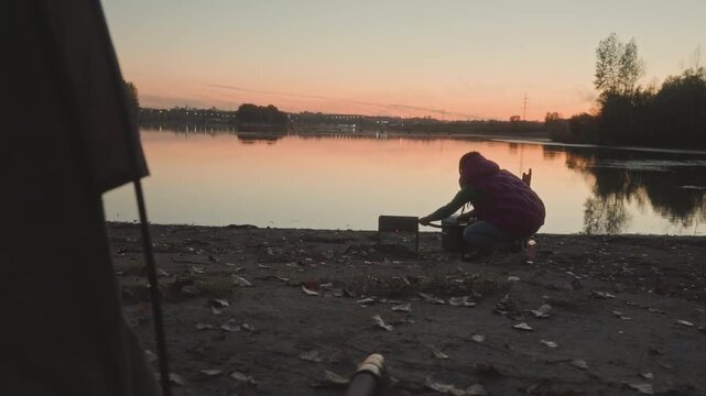 Sunset lake angler setting up fishing gear on rocky shore, twilight glow and water reflections, person crouched arranging rod and tackle boxes, portable stove and bucket nearby, calm breeze, focused
