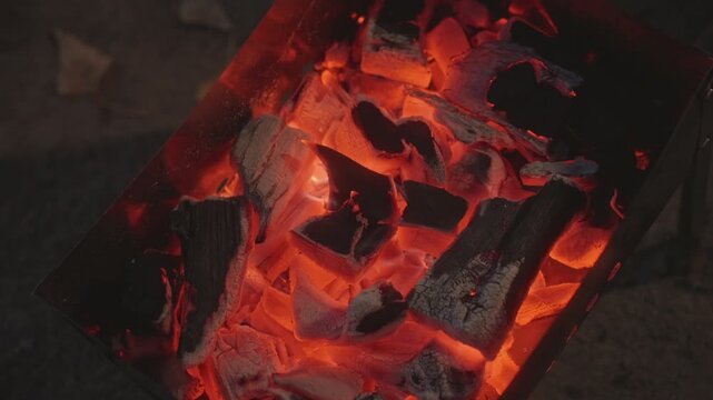 Glowing coals in campfire pit, molten embers and charcoal pieces radiating deep orange heat, subtle smoke rising, occasional sparks, cozy evening vibe ideal for camping scenes, cooking over grill,