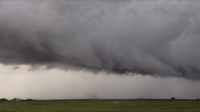 A skinny rope tornado writhes around in the sky and reaches to the ground as dark storm clouds brew over a field.