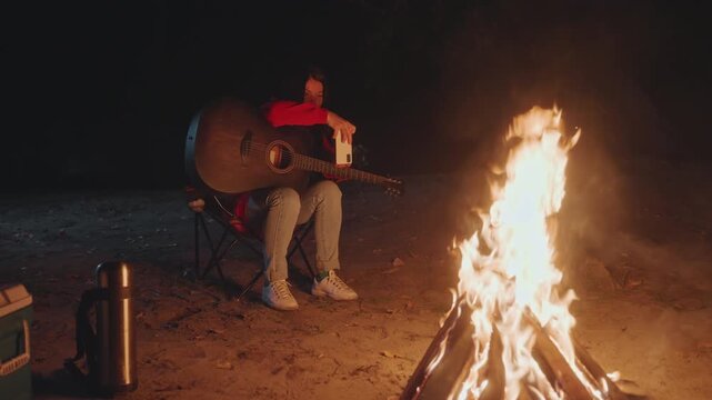 Night campfire guitar session asian male solo camper strums acoustic guitar on folding chair beside roaring bonfire, warm firelight, cooler and thermos nearby, reflective mood, gentle strumming,
