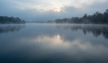 Fototapeta premium Misty morning drifts over calm lake surface. Soft light glows through hazy sky above trees. Reflections ripple gently across still water. Fog curls low near shoreline, veiling distant shore