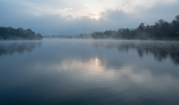 Misty morning drifts over calm lake surface. Soft light glows through hazy sky above trees. Reflections ripple gently across still water. Fog curls low near shoreline, veiling distant shore